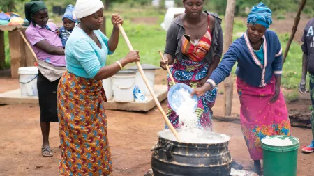 Three women surround a large metal pot. Two are stirring while one is throwing a bowl of rice into the hot water.