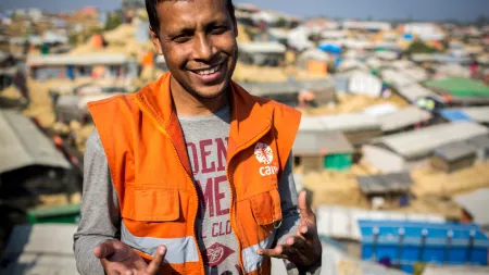 A man wearing an orange CARE vest smiles and gestures towards the camera. Behind him, a large town is visible.