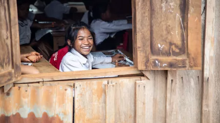 A Cambodian girl setting at a desk smiles widely. The picture is taken from outside, and she is by an open window.