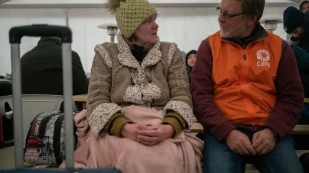 A Ukrainian woman, dressed warmly and covered by a light pink blanket, sits and talks with a man wearing an orange CARE vest.