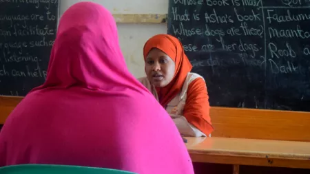 Somalia_Female teacher in CARE vest sitting behind desk talking to lady in pink niqab