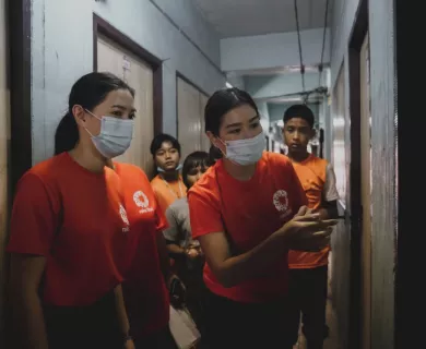 Two Thai women, each wearing a bright orange CARE t-shirt and a light blue surgical mask, lead a group of children through a hallway.