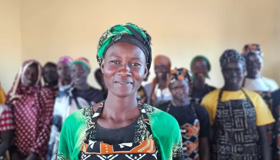 Woman in the forefront wearing apron, t-shirt and head scarf with patterns, with a group of women in the backgrounf