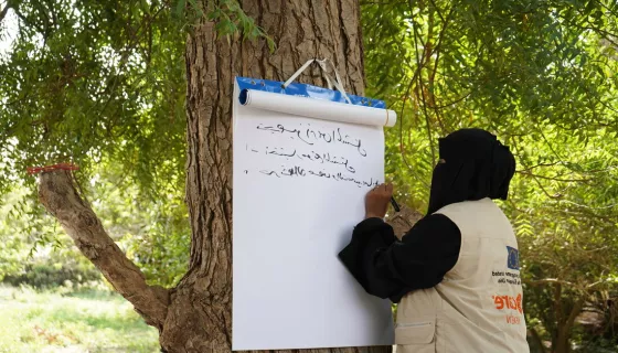 Woman wearing black head scarf and face cover writing on large block of paper hanging on a tree