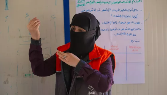 Woman wearing black head scarf and face cover with gesticulating in a room with large information paper up on the wall.