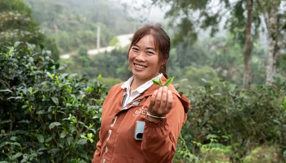 Woman wearing orange jacket, holding a tea leaf and smiling at the camera, while standing in a green area
