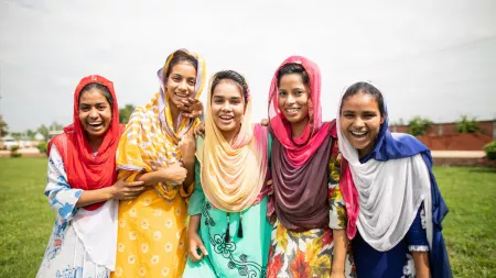 A group of five young women smile and laugh together while standing in a field. They are all wearing brightly colored dresses and head scarves.