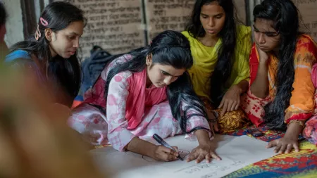 A young woman sits on the ground and writes on a large sheet of white paper in black marker. A few other young women sit on either side of her, watching intently as she writes.