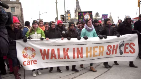 A group of young women smile and march while carrying a long white banner that says, "CARE Walk In Her Shoes." It's an overcast day and they are wearing coats, gloves, and hats.