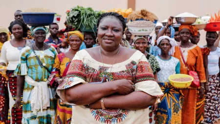 A woman stands in center in front of a large group of women carrying bowls and baskets of fruits, veggies, and grains.