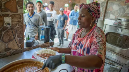 A woman spoons beans and rice onto plates while a long line of people wait outside.