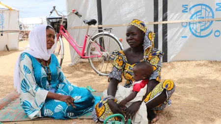 Two women sit in sand in front of a large, white tent. One of them is holding a small baby in her lap.