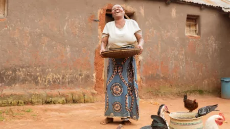 A woman smiles and laughs while holding a flat circular basket full of seeds. Beside her a group of chickens pecking.