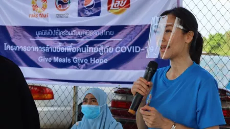 A woman wearing a protective face shield speaks into a microphone. Behind her is a sign that says, "Give meals give hope."