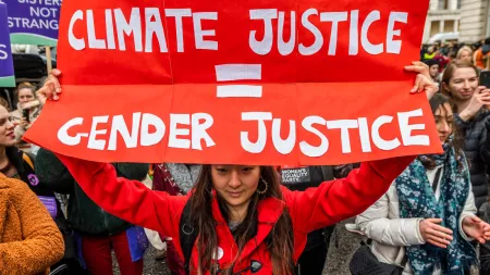 A woman holds a bright red handmade sign over her head. The sign reads, "CLIMATE JUSTICE = GENDER JUSTICE."