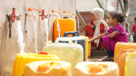 Two young girls fill up large yellow jugs at a tap water station.