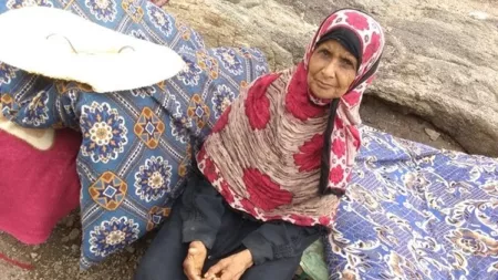 An older Yemeni woman sits on the ground, leaning against some fabric.