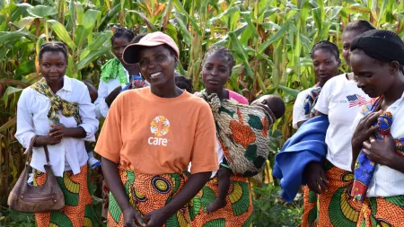 A group of women, some of them carrying babies, stand together in front of a corn field. The woman in the center is wearing a bright orange CARE t-shirt and an orange and green patterned skirt.