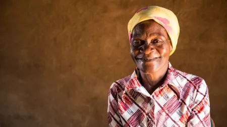 A portrait of a Zimbabwean woman. She is smiling kindly in front of a dark brown wall, and is wearing a patterned blouse and a yellow headwrap.