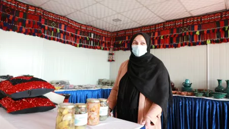 A woman wearing a head scarf and surgical face mask stands at a table with jarred and canned goods.