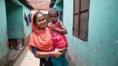 A Bangladeshi woman smiles while holding a child in her arms. She is standing in an alleyway between two buildings painted a cheerful teal blue. In the background, a few colorful items of clothing are hanging from a clothing line.