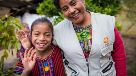 A Guatemalan woman and girl smile and wave. The woman is wearing a white jacket with a CARE logo embroidered above the pocket.