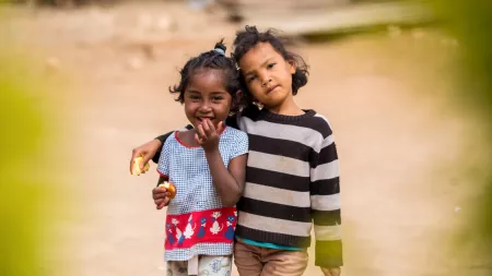 Two girls stand in the middle of the road, each holding food in one hand. One of the girls has her arm around the other's shoulders and is resting her head on hers. The second girl is bringing up her hand up to her mouth as she smiles.