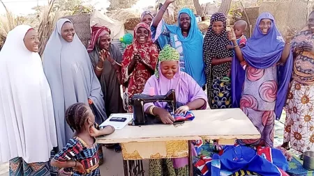 A woman sits at a desk outside and sews a garment with a sewing machine. She is surrounded by women smiling and cheering her on.