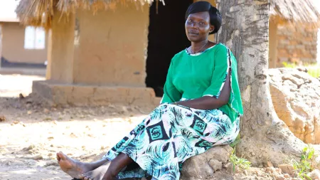 A Ugandan woman wearing a bright green top and a patterned skirt sits on the ground and leans against a tree.