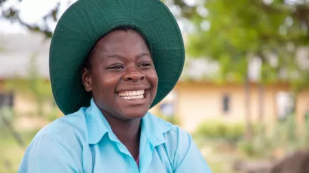 A woman wearing a bright blue collared shirt and green sun hat smiles widely.