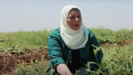 A Syrian woman wearing an emerald green shirt and a white head scarf sits in a field and talks.
