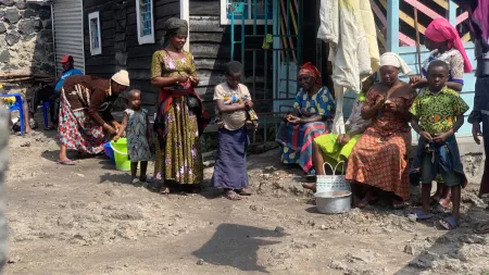 Woman standing between two children in front of other women sitting outside a house