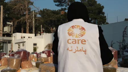 CARE staff's back wearing CARE's jacket and black clothes in a field with food bags for distribution
