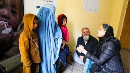Woman in burqa with her two children speaking with woman kneeling holding her hands and man sitting in the floor in the background