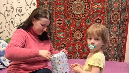 Ukraine_Mother unwrapping gift for little girl on bed