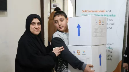 Boy carrying boxes with CARE's logo and woman on the left wearing black headscarf and touching boy's arm