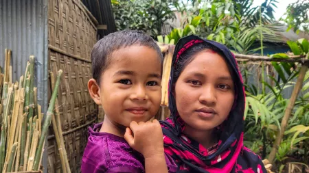 Bangladesh_Woman with black scarf holding daughter with purple dress on in front of shack