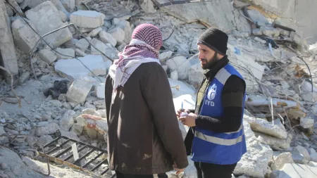 Syria_Man wearing IYD vest talking to Syrian man with red and white scarf in front of rubble
