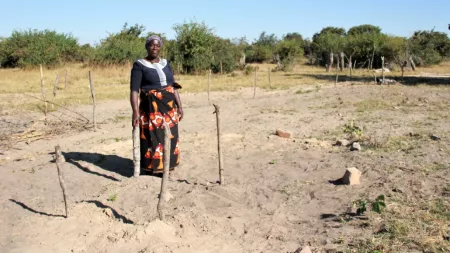 Zambia_Woman standing in sandy area with four sticks surrounding dug hole