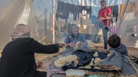 A gray haired man holding bread sitting on the floor of a plastic tent, a woman to his left handing in more bread, a boy to the left, and a man stading behind 