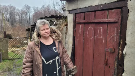 Woman with grey hair and pink coat in front of wooden door with inscription made with white chalk