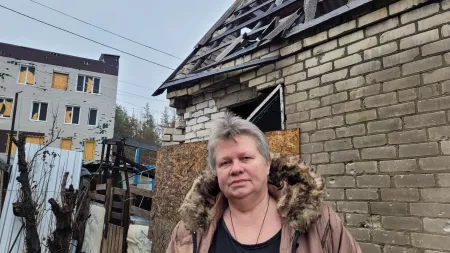 Woman with grey hair and pink coat in front of house with destroyed roof