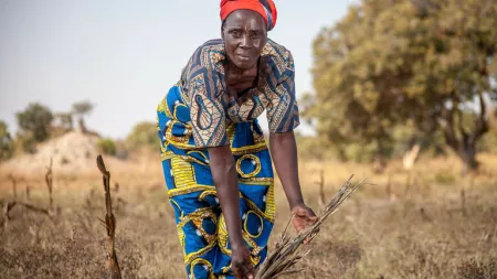 A woman wearing blue and yellow patterned clothes shows her withered crops in the field. Dried grass and tress in the background.