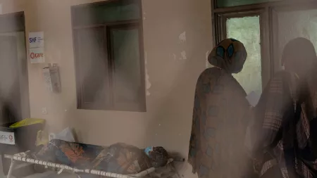 A person lying on a cot, covered with a patterned blanket, while two women in patterned shawls stand nearby in a dimly lit room with CARE and SHF signage.