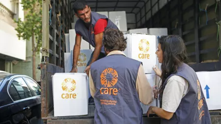 CARE aid workers load boxes marked with CARE logo onto a truck. Three people visible, wearing CARE vests. Scene shows humanitarian supply distribution in progress.