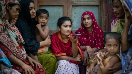 Group of South Asian women and children sitting together. Women wear colorful headscarves. Young woman in red holds smartphone. 