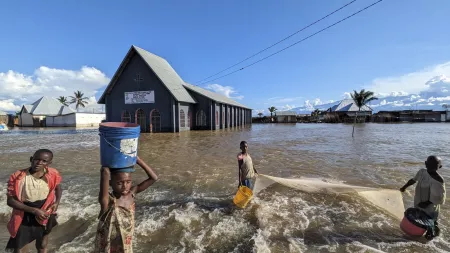 Children carry water buckets while wading through floods near a submerged church in Gatumba, daily life persisting amid disaster.