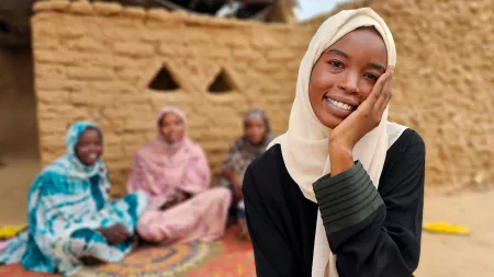 Woman beige scarf smiling to the camera and leaning her hand on her face with three women blurred in the background.