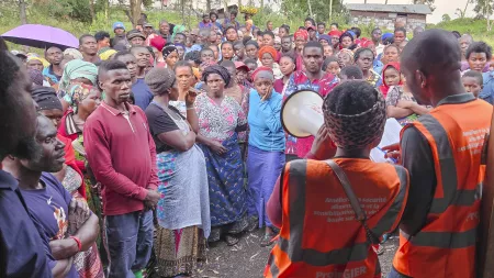A diverse crowd listens to aid workers in orange vests at an outdoor gathering. People wear colorful clothes while workers use megaphone to address them.