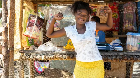 Young woman wearing light t-shirt and yellow skirt, showing her strong arms, while standing in front of her small shop